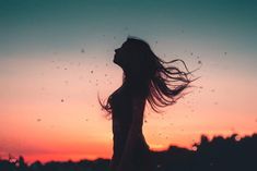 the silhouette of a woman with her hair blowing in the wind as the sun sets