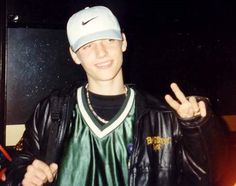 a young man in a leather jacket and baseball cap making the peace sign with his hand