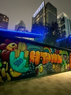 a wall covered in lots of colorful graffiti next to tall buildings at night with lights on