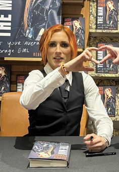 a woman with red hair sitting at a table in front of books and holding her finger up