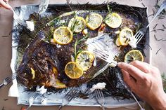 a person holding a fork and knife over a fish on a tray with lemons