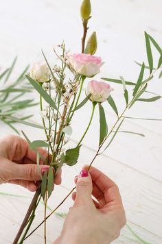 two hands are arranging flowers on a white wooden surface with green leaves and stems in the foreground