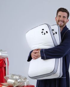 a man in pajamas holding a pillow with faces on it and wrapped presents behind him