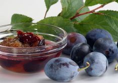 plums and blueberry jam in a glass bowl on a white surface next to green leaves