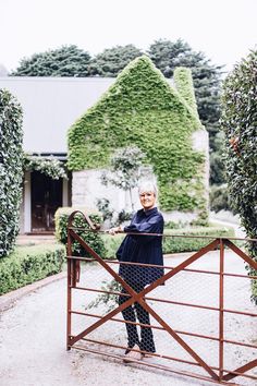 an older woman standing behind a gate in front of a house with ivy growing on it