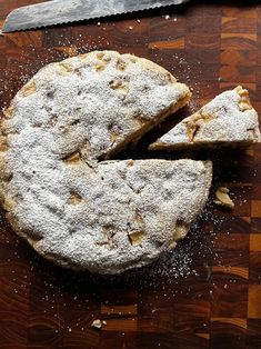 a wooden cutting board topped with a pie covered in powdered sugar next to a knife