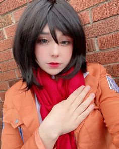 a woman with black hair wearing an orange jacket and red scarf standing in front of a brick wall