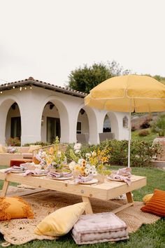 a picnic table with yellow umbrellas and pillows on the grass in front of a house