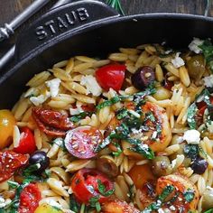 a skillet filled with pasta and vegetables on top of a wooden table next to a fork