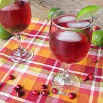 two glasses of cranberry punch sit on a table with limes and cherries