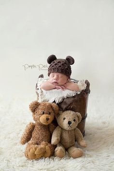 a baby in a basket with two teddy bears on the floor next to it is wearing a hat