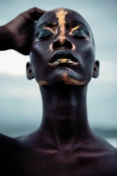 a woman with gold painted on her face and hands holding her hair up to her head