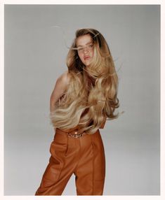 a woman with long blonde hair standing in front of a gray background wearing brown pants