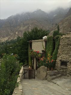 a stone building with flowers growing on the side and mountains in the background, near a gate