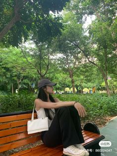 a woman sitting on top of a wooden bench in a park next to lush green trees