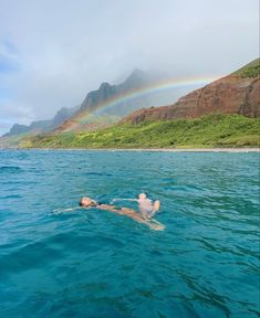 a man swimming in the ocean with a rainbow above him and mountains in the background