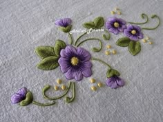 purple flowers and green leaves are embroidered onto a white tablecloth with yellow thread on it