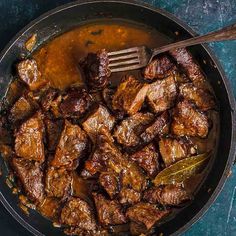 a pan filled with meat and sauce on top of a blue countertop next to a wooden spoon