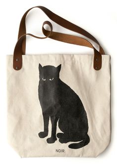 a black cat sitting on top of a white tote bag with brown handles and straps