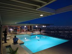 an indoor swimming pool at night with people standing around it and the city lights in the background