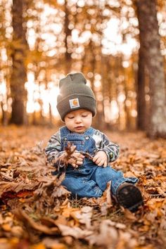 a little boy sitting in the leaves with a hat on