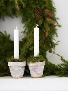 two white candles are placed in small pots with moss and pine cones on the mantle