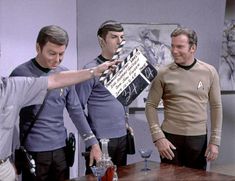 three men in star trek uniforms standing around a table and pointing at something on it