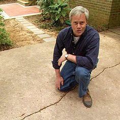 a man squatting down on the ground in front of a brick building with trees and bushes behind him