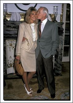 an older man and woman standing next to each other in front of a wall with a clock on it