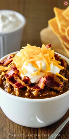 a white bowl filled with chili and cheese next to some tortilla chips on a wooden table