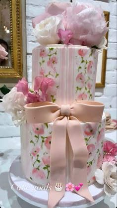 a three tiered cake with pink and white flowers on the top, decorated with bows