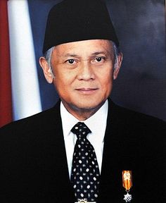 an older man wearing a suit and tie with a medal on his lapel, standing in front of a flag