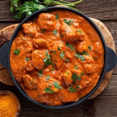 a pan filled with chicken curry next to spices and an orange sauce on a wooden board