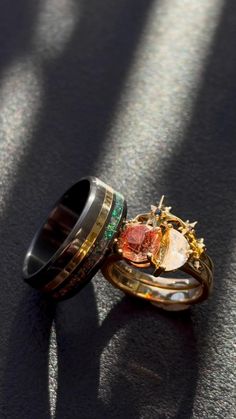 A matching wedding ring set from Staghead Designs sits on a black surface with rays of sun beaming in. On the left is our "Cosmo" black zirconium wedding band with an offset channel of black opal & another channel with a yellow gold inlay. On the right is the "Sidra" engagement ring set in 14K yellow gold with a thick shank featuring diamond accented star details, a toi et moi type design with a round Oregon sunstone & crescent moon moissanite, & matching starry tracer with sapphire accents. Celestial Wedding Ring, Moon Dancing, Whiskey Barrel Wedding Ring, Sunstone Engagement Ring, Engament Rings, Antler Wedding Rings, Men's Wedding Rings, Handcrafted Engagement Ring, Wood Wedding Ring