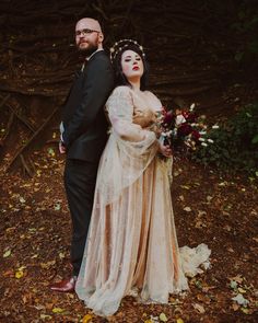 a man and woman standing next to each other in front of trees with leaves on the ground