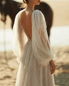 a woman in a white dress standing next to a brown horse on the beach with her back turned