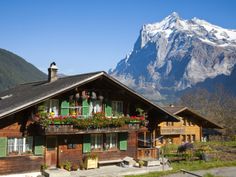 a wooden house with green shutters and flowers on the balcony in front of mountains