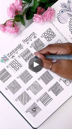 someone is drawing designs on a notebook with a pen and some pink flowers in the background