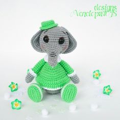 a crocheted elephant in a green dress sitting on the ground with flowers around it