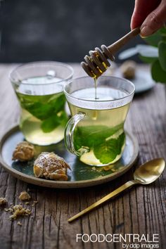 two cups filled with green tea on top of a wooden table next to gold spoons