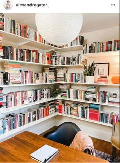 a room filled with lots of books next to a wooden table and white ceiling light