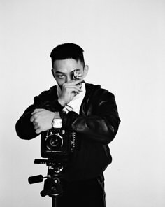 black and white photograph of man holding camera with hand over mouth, looking at camera