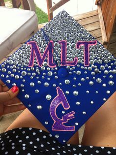 a blue graduation cap decorated with pink letters and silver dots on it, sitting in front of a woman's legs