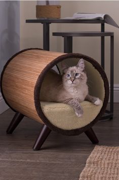 a cat is sitting in a wooden barrel shaped bed on the floor next to a table