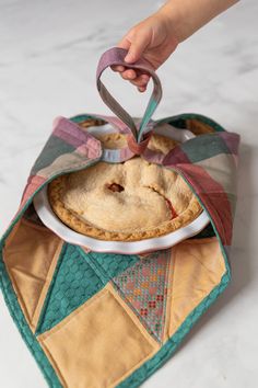 a hand holding a pie in a quilted pot holder on top of a table