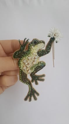 a hand holding a beaded frog brooch in it's right hand, with a flower on its left side