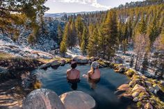 two people sitting in the middle of a pool surrounded by rocks and pine trees, with snow on the ground