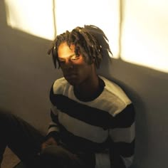 a man with dreadlocks sitting on the floor in front of a wall and sunlight coming through the window