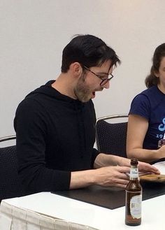 two people sitting at a table with food and drinks in front of them, one person holding a bottle