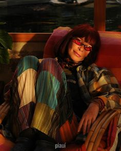 a woman with red hair and glasses sitting on a chair in the sun wearing colorful clothing